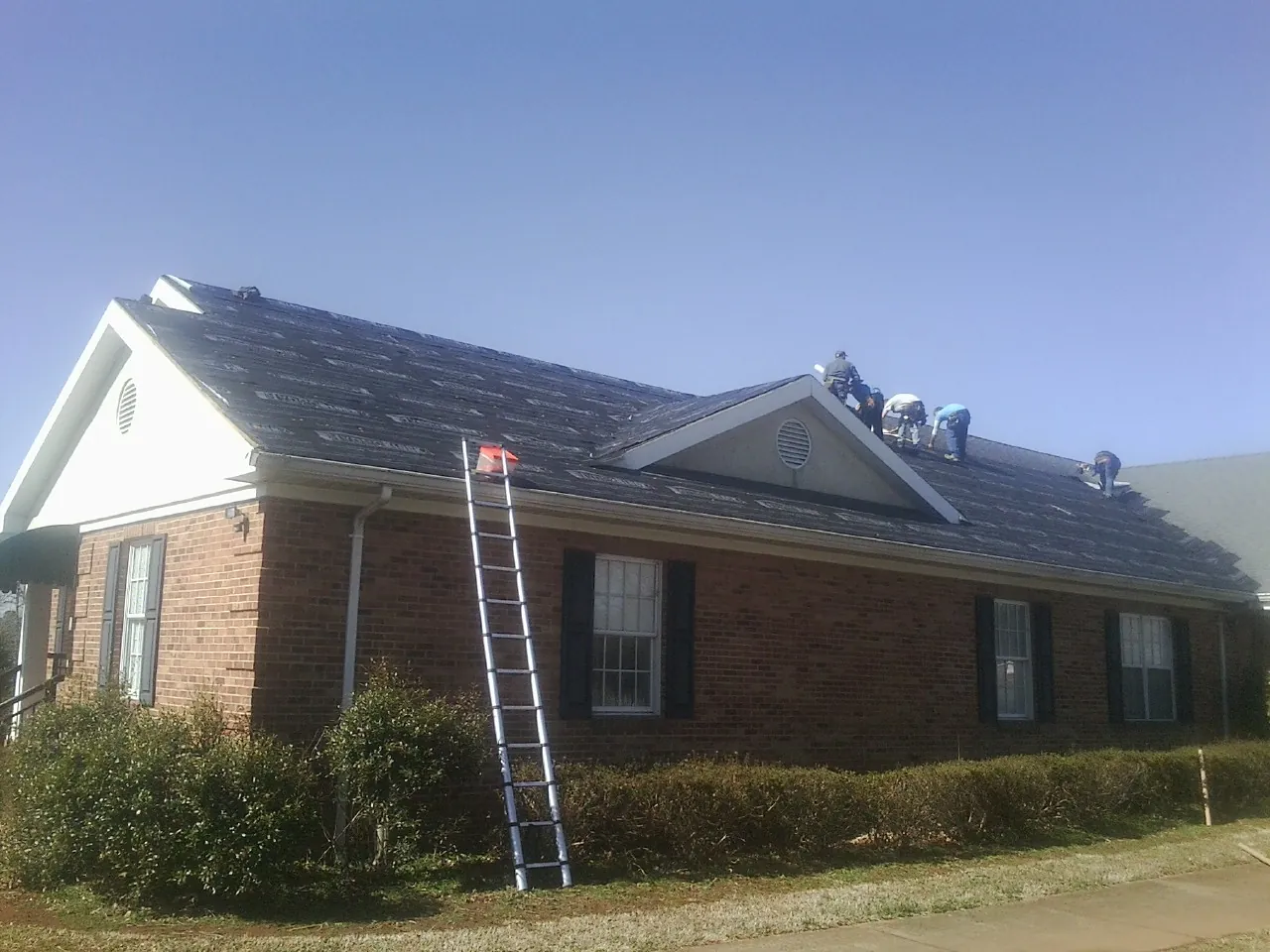 Crew tearing shingles off a brick ranch roof with a ladder set — Emergency Roof Repair work by our crew in Minier